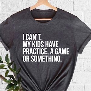 May include: Dark gray t-shirt with the text "I CAN'T. MY KIDS HAVE PRACTICE, A GAME OR SOMETHING." in white. The shirt is displayed on a wooden hanger against a white brick wall, with green plants in the foreground.