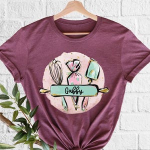 May include: Burgundy t-shirt featuring a baking-themed graphic. The design includes a whisk, piping bags, and a spatula, with the name "Gabby" on a rolling pin. The graphic is set against a pink background.