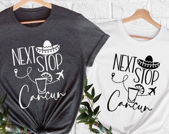 May include: Two t-shirts, one dark gray and one white, each with the phrase "NEXT STOP Cancun" in black script. The design includes a sombrero, a cocktail, and an airplane. The shirts are hanging on wooden hangers.