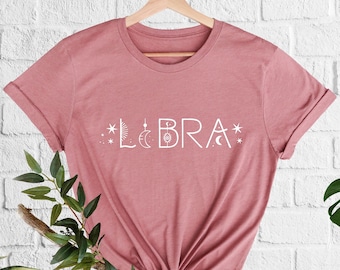 May include: A dusty rose t-shirt with a white graphic design that says "LIBRA" with stars and moon symbols.