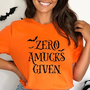 May include: A bright orange short-sleeved t-shirt featuring the black text "ZERO AMUCKS GIVEN" printed on the front, accompanied by small black bat silhouettes. The shirt is paired with blue denim jeans that have a distressed, ripped detail.