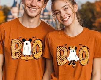 May include: Two orange short-sleeve t-shirts featuring a graphic print of the word BOO. The letter O is replaced by a white ghost character with mouse ears. The letter B has a black polka dot pattern, and the letter O has a pumpkin pattern.
