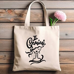 May include: A white canvas tote bag with black handles and a black graphic of a woman's face with the text "Gemini" above her head.