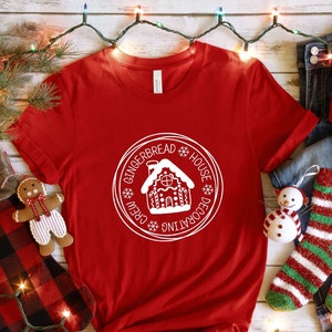 May include: Red t-shirt with a white graphic of a gingerbread house and the text "Gingerbread House Decorating Crew".