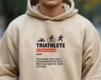May include: A beige hoodie with a black graphic design that says "TRIATHLETE" and defines the word as "someone who can't comprehend the fact that one sport is hard enough". The graphic also includes icons of a swimmer, a cyclist, and a runner.