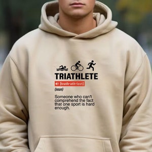 以下が含まれることがあります： トライアスロンの言葉が「トライアスロン」と定義され、「1つのスポーツが十分に難しいという事実を理解できない人」と定義された、ベージュのフード付きスウェットシャツ。グラフィックには、スイマー、サイクリスト、ランナーのアイコンも含まれています。