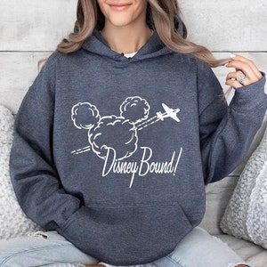 Puede incluir: Una sudadera con capucha azul oscuro con un gr&aacute;fico blanco de la cabeza de Mickey Mouse en nubes con un avi&oacute;n volando por encima. El texto "Disney Bound!" est&aacute; debajo del gr&aacute;fico.