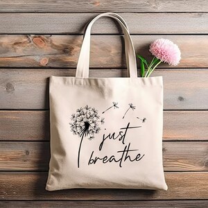 May include: A white canvas tote bag with black text that reads "just breathe" and a black dandelion graphic with seeds blowing in the wind.