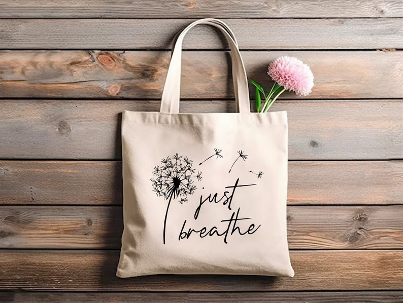 May include: A white canvas tote bag with black text that reads "just breathe" and a black dandelion graphic with seeds blowing in the wind.