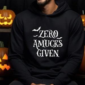 May include: A black hooded sweatshirt featuring white text that reads ZERO AMUCKS GIVEN, accompanied by small white bat silhouettes. The hoodie has long sleeves, a front pocket, and a hood, displaying a graphic design on the chest area.