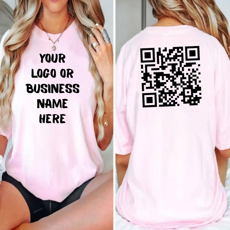 May include: A light pink t-shirt with the text "YOUR LOGO OR BUSINESS NAME HERE" printed in black on the front. The back of the shirt features a black QR code. The shirt has short sleeves.