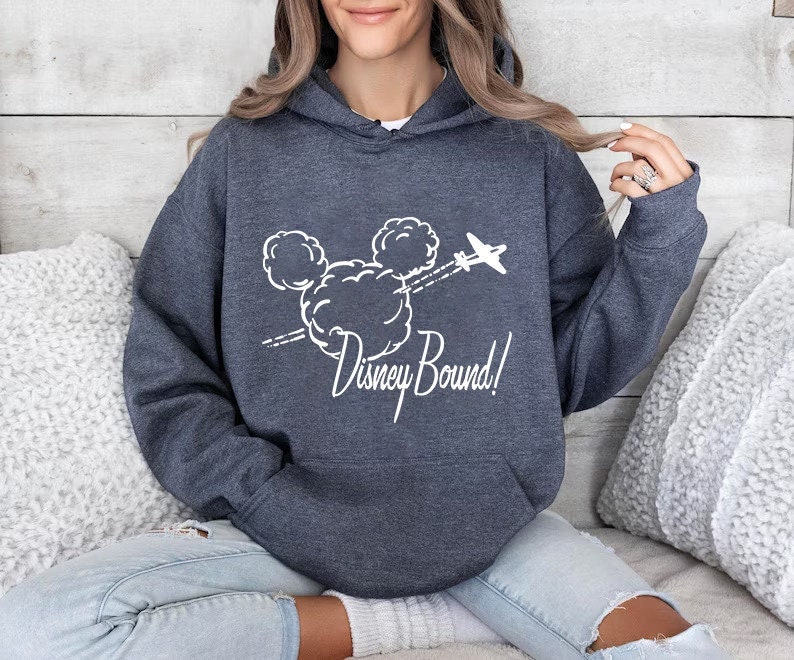 Puede incluir: Una sudadera con capucha azul oscuro con un gr&aacute;fico blanco de la cabeza de Mickey Mouse en nubes con un avi&oacute;n volando por encima. El texto "Disney Bound!" est&aacute; debajo del gr&aacute;fico.