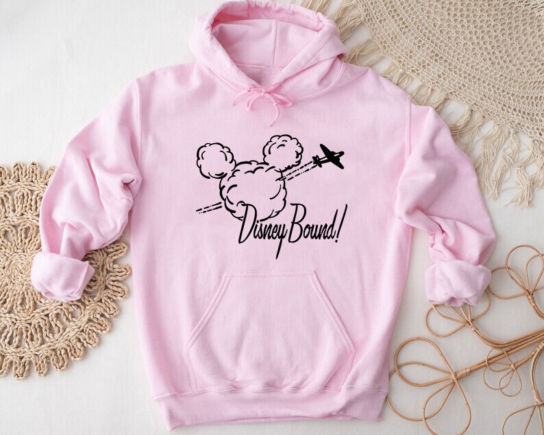 Puede incluir: Sudadera rosa con capucha con un dise&ntilde;o negro de cabeza de Mickey Mouse y un avi&oacute;n volando a trav&eacute;s de una nube. El texto "Disney Bound!" est&aacute; impreso debajo del dise&ntilde;o.