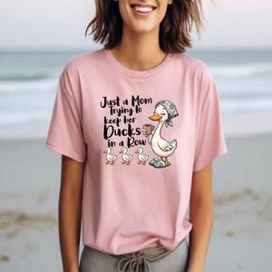 以下が含まれることがあります： 「Just a Mom Trying to Keep Her Ducks in a Row」の文字が入ったピンク色のTシャツ。頭にスカーフを巻いてコーヒーカップを持った漫画の鴨と、その下に3羽のアヒルの子がいるグラフィック。