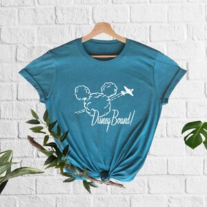 Peut inclure: Un t-shirt bleu turquoise avec un graphique blanc repr&eacute;sentant la t&ecirc;te de Mickey Mouse faite de nuages avec un avion volant au-dessus. Le texte "Disney Bound!" est sous le graphique.