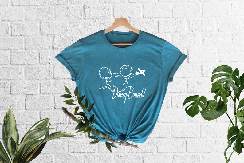 Peut inclure: Un t-shirt bleu turquoise avec un graphique blanc repr&eacute;sentant la t&ecirc;te de Mickey Mouse faite de nuages avec un avion volant au-dessus. Le texte "Disney Bound!" est sous le graphique.