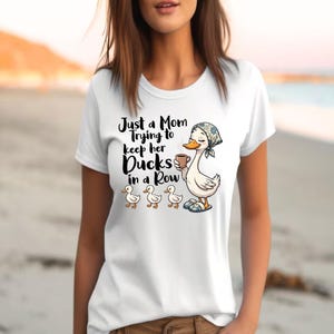 以下が含まれることがあります： 「Just a Mom Trying to Keep Her Ducks in a Row」の文字が入った白いTシャツ。イラストは、コーヒーカップを持ち、スカーフをかぶり、3羽のアヒルの隣に立っている漫画のアヒルが特徴です。