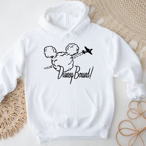 Puede incluir: Sudadera con capucha blanca con un gr&aacute;fico negro de las orejas de Mickey Mouse con un avi&oacute;n volando a trav&eacute;s de ellas y el texto "Disney Bound!"