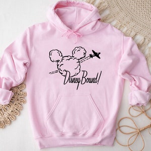 Puede incluir: Sudadera rosa con capucha con un dise&ntilde;o negro de cabeza de Mickey Mouse y un avi&oacute;n volando a trav&eacute;s de una nube. El texto "Disney Bound!" est&aacute; impreso debajo del dise&ntilde;o.