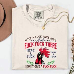 May include: Cream-colored t-shirt with a graphic of a rooster and the text "WITH A FUCK FUCK HERE And a FUCK FUCK THERE HERE A FUCK THERE A FUCK I DON'T GIVE A FUCK FUCK". A gold necklace with a star pendant is draped on the shirt.