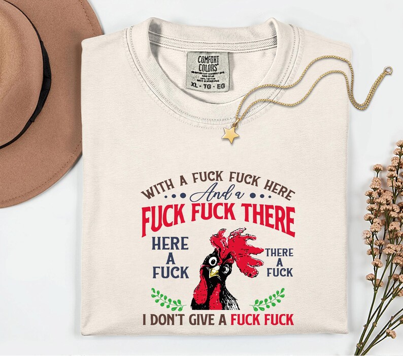 May include: Cream-colored t-shirt with a graphic of a rooster and the text "WITH A FUCK FUCK HERE And a FUCK FUCK THERE HERE A FUCK THERE A FUCK I DON'T GIVE A FUCK FUCK". A gold necklace with a star pendant is draped on the shirt.
