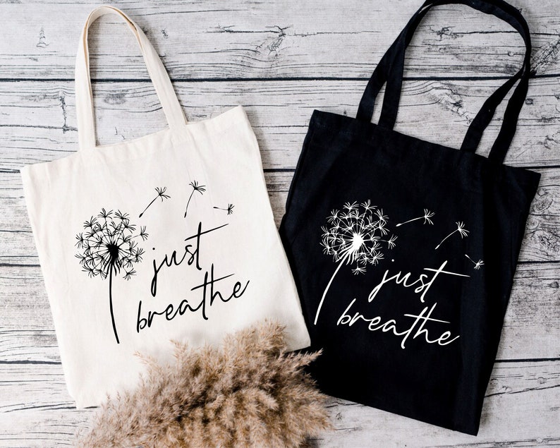 May include: Two tote bags with the text "just breathe" and a dandelion graphic. One bag is white with black text and graphic, the other is black with white text and graphic.
