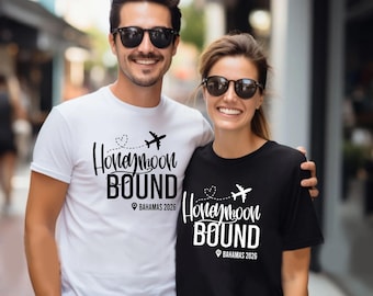 Może przedstawiać: Biała i czarna koszulka z napisem "Honeymoon Bound", grafiką samolotu i serca. Zawiera również tekst "Bahamas 2026". Koszulki nosi uśmiechnięta para.
