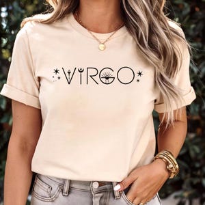 May include: A beige t-shirt with the word "VIRGO" in black, featuring a celestial design. The design includes stars, a crescent moon, and a sun-like symbol. The shirt has short sleeves and a round neckline.