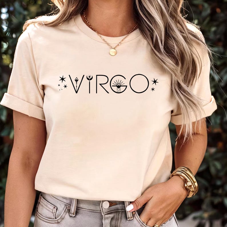 May include: A beige t-shirt with the word "VIRGO" in black, featuring a celestial design. The design includes stars, a crescent moon, and a sun-like symbol. The shirt has short sleeves and a round neckline.
