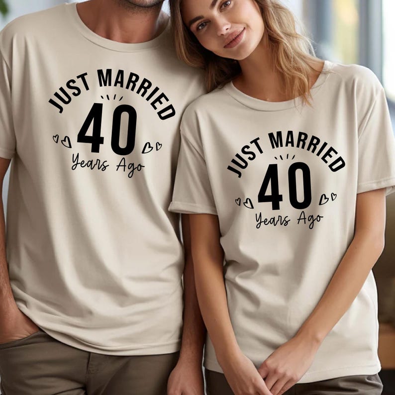 May include: Two beige t-shirts with the text "JUST MARRIED 40 Years Ago" in black. The number 40 is large and centered, with small heart details. The shirts are short-sleeved.