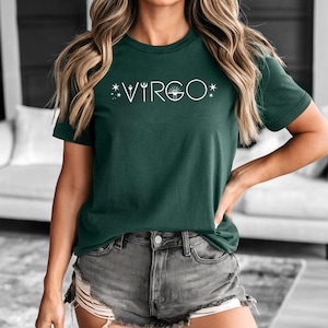 May include: Dark green t-shirt with the word "VIRGO" in white, along with star and sunburst graphics. The shirt is a crew neck and short-sleeved. The model is wearing gray denim shorts.