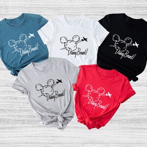 Peut inclure: Cinq t-shirts de diff&eacute;rentes couleurs avec un motif de t&ecirc;te de Mickey Mouse dans les nuages avec un avion qui vole au-dessus. Le texte "Disney Bound!" est imprim&eacute; sur chaque t-shirt.