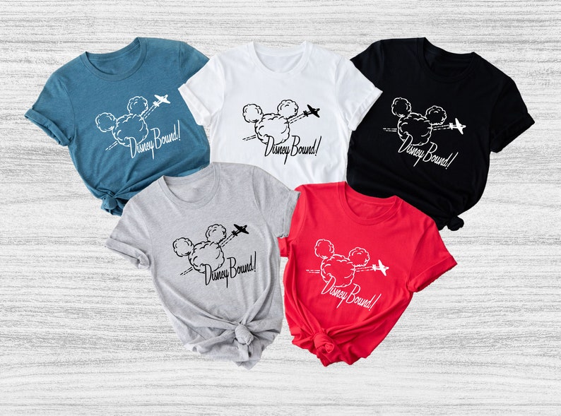 Peut inclure: Cinq t-shirts de diff&eacute;rentes couleurs avec un motif de t&ecirc;te de Mickey Mouse dans les nuages avec un avion qui vole au-dessus. Le texte "Disney Bound!" est imprim&eacute; sur chaque t-shirt.