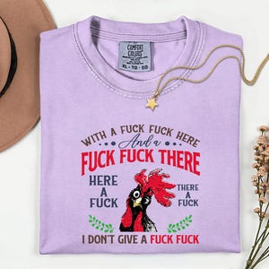 May include: A light purple t-shirt with the text "WITH A FUCK FUCK HERE And a FUCK FUCK THERE HERE A FUCK THERE A FUCK I DON'T GIVE A FUCK FUCK" and a rooster graphic. A gold necklace with a star pendant is draped across the shirt.
