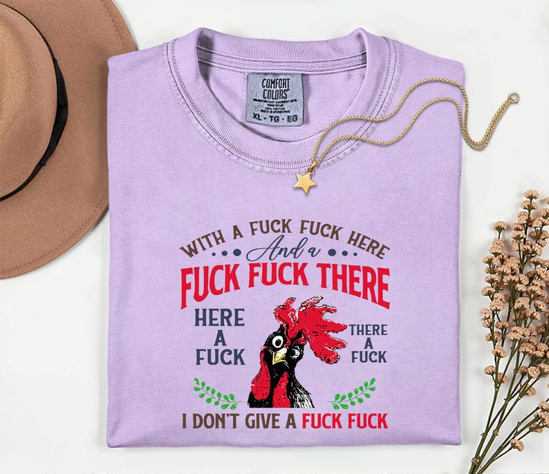 May include: A light purple t-shirt with the text "WITH A FUCK FUCK HERE And a FUCK FUCK THERE HERE A FUCK THERE A FUCK I DON'T GIVE A FUCK FUCK" and a rooster graphic. A gold necklace with a star pendant is draped across the shirt.