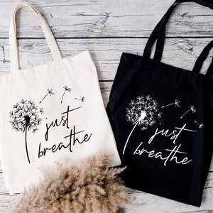 May include: Two tote bags with the text "just breathe" and a dandelion graphic. One bag is white with black text and graphic, the other is black with white text and graphic.