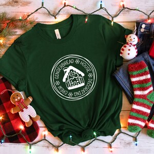 May include: A dark green short-sleeve t-shirt featuring a white circular graphic with a gingerbread house illustration and the text "GINGERBREAD HOUSE DECORATING CREW" surrounded by snowflake icons, centered on the chest of the garment.