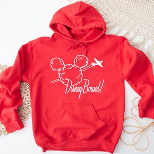 Puede incluir: Sudadera roja con capucha con un gr&aacute;fico blanco de la cabeza de Mickey Mouse en nubes con un avi&oacute;n volando por encima. El texto "Disney Bound!" est&aacute; debajo del gr&aacute;fico.