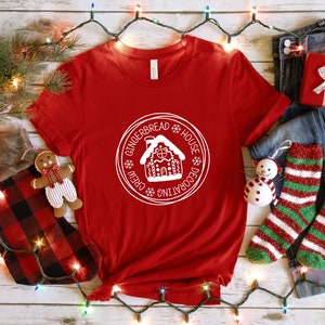 May include: A red short-sleeved t-shirt featuring a white circular graphic in the center. The graphic depicts a gingerbread house surrounded by the text "GINGERBREAD HOUSE DECORATING CREW" and small snowflake icons. The shirt has a crew neckline.