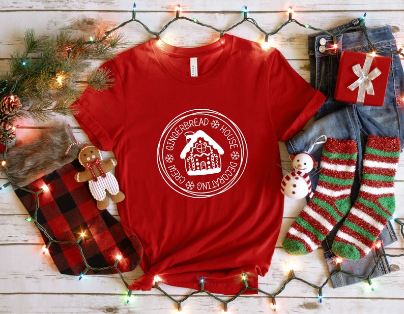 May include: A red short-sleeved t-shirt featuring a white circular graphic in the center. The graphic depicts a gingerbread house surrounded by the text "GINGERBREAD HOUSE DECORATING CREW" and small snowflake icons. The shirt has a crew neckline.