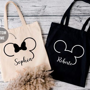May include: Two tote bags with custom names. One bag is beige with a black Minnie Mouse silhouette and the name "Sophia". The other bag is black with a white Mickey Mouse silhouette and the name "Roberto".