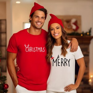 May include: A couple wearing matching red and white Christmas t-shirts and red elf hats. The red shirt says "Christmas" in white script. The white shirt says "Merry" in black script.