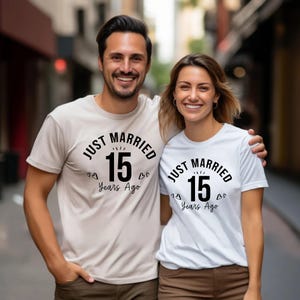 May include: A couple wearing matching t-shirts with the text "JUST MARRIED 15 Years Ago" in black. The man wears a beige shirt, and the woman wears a white shirt. They are smiling and embracing.