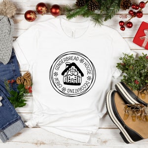 May include: A white t-shirt featuring a black circular graphic of a gingerbread house with the text "GINGERBREAD HOUSE DECORATING CREW" and snowflake icons. The design is centered on the chest of the shirt, which is displayed on a flat surface.