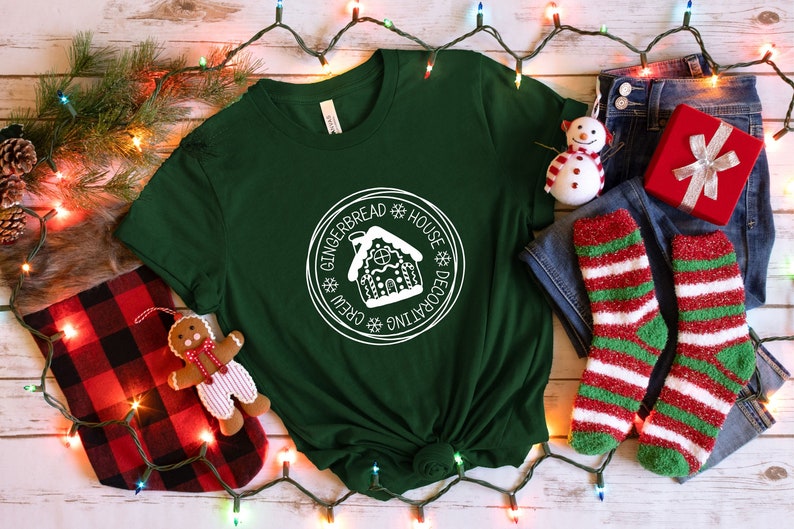 May include: A dark green short-sleeve t-shirt featuring a white circular graphic with a gingerbread house illustration and the text "GINGERBREAD HOUSE DECORATING CREW" surrounded by small snowflake icons, centered on the chest of the garment.