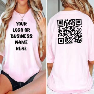 May include: A light pink t-shirt with the text "YOUR LOGO OR BUSINESS NAME HERE" printed in black on the front. The back of the shirt features a black QR code. The shirt has short sleeves.