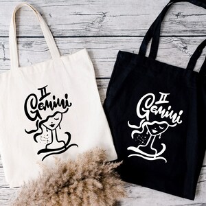 May include: Two tote bags with the Gemini zodiac sign and a stylized illustration of a woman's face. One bag is white with black text and illustration, the other is black with white text and illustration.