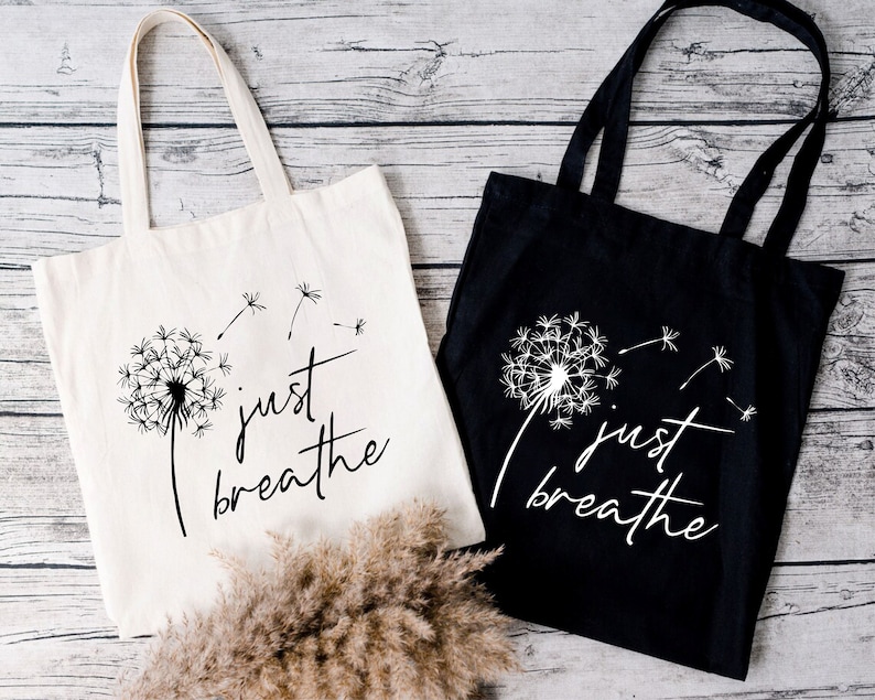 May include: Two tote bags with the text "just breathe" and a dandelion graphic. One bag is white with black text and graphic, the other is black with white text and graphic.