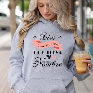 May include: A gray hooded sweatshirt with a black and pink graphic design that reads "Dios tiene un plan que lleva tu nombre" with the text "Proverbios 16:9" underneath.
