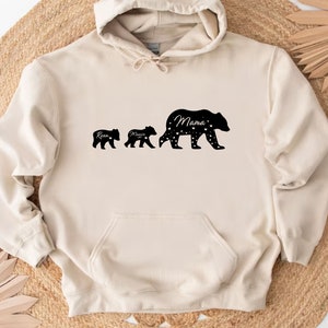 May include: A cream-colored hoodie with a black silhouette of a mama bear and two cubs. The mama bear has the word "Mama" written on her back. The cubs are labeled "Ryan" and "Maya".